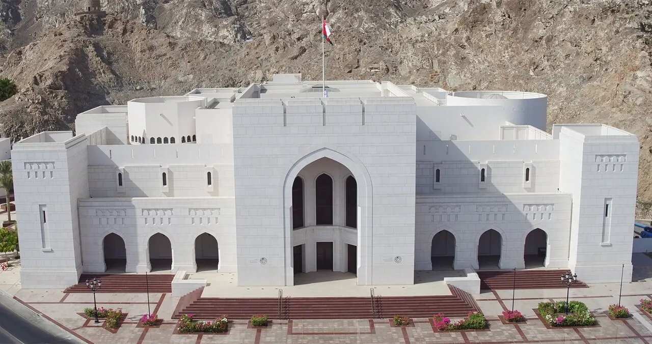 A white symmetrical building with a large central arch and smaller arches on each side, a flag on top and a rocky hill behind.