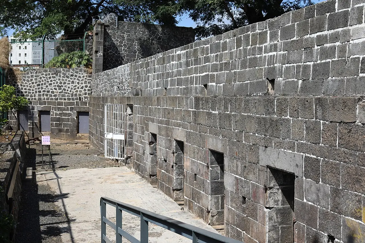 Os muros de pedra basáltica escura do Aapravasi Ghat, em Port Louis, com uma passagem estreita entre eles e uma grade de proteção.