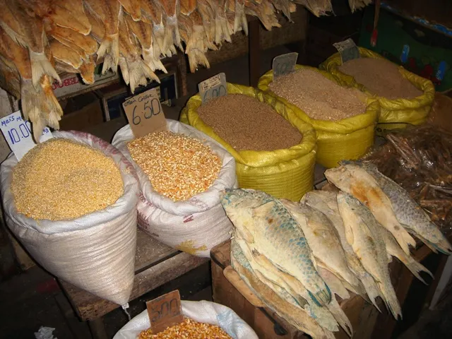 Uma banca do Central Market de Port Louis com sacos abertos de grãos e lentilhas, peixes secos e plaquinhas de preço.