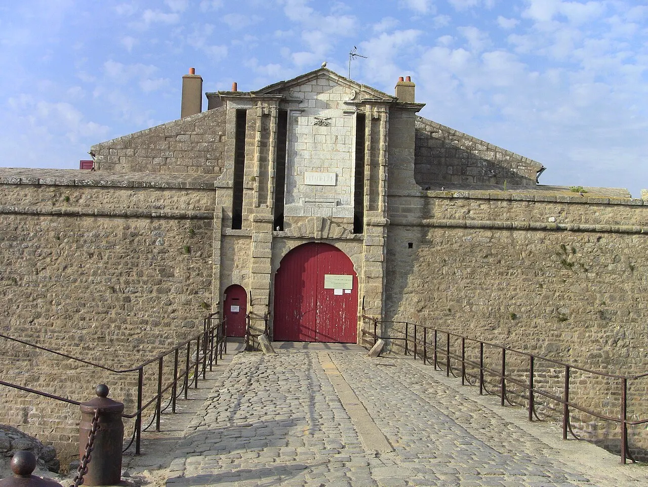 A entrada do Fort Adelaide, em Port Louis, com muralha de pedra cinza, portão vermelho em arco e uma passarela com corrimão.