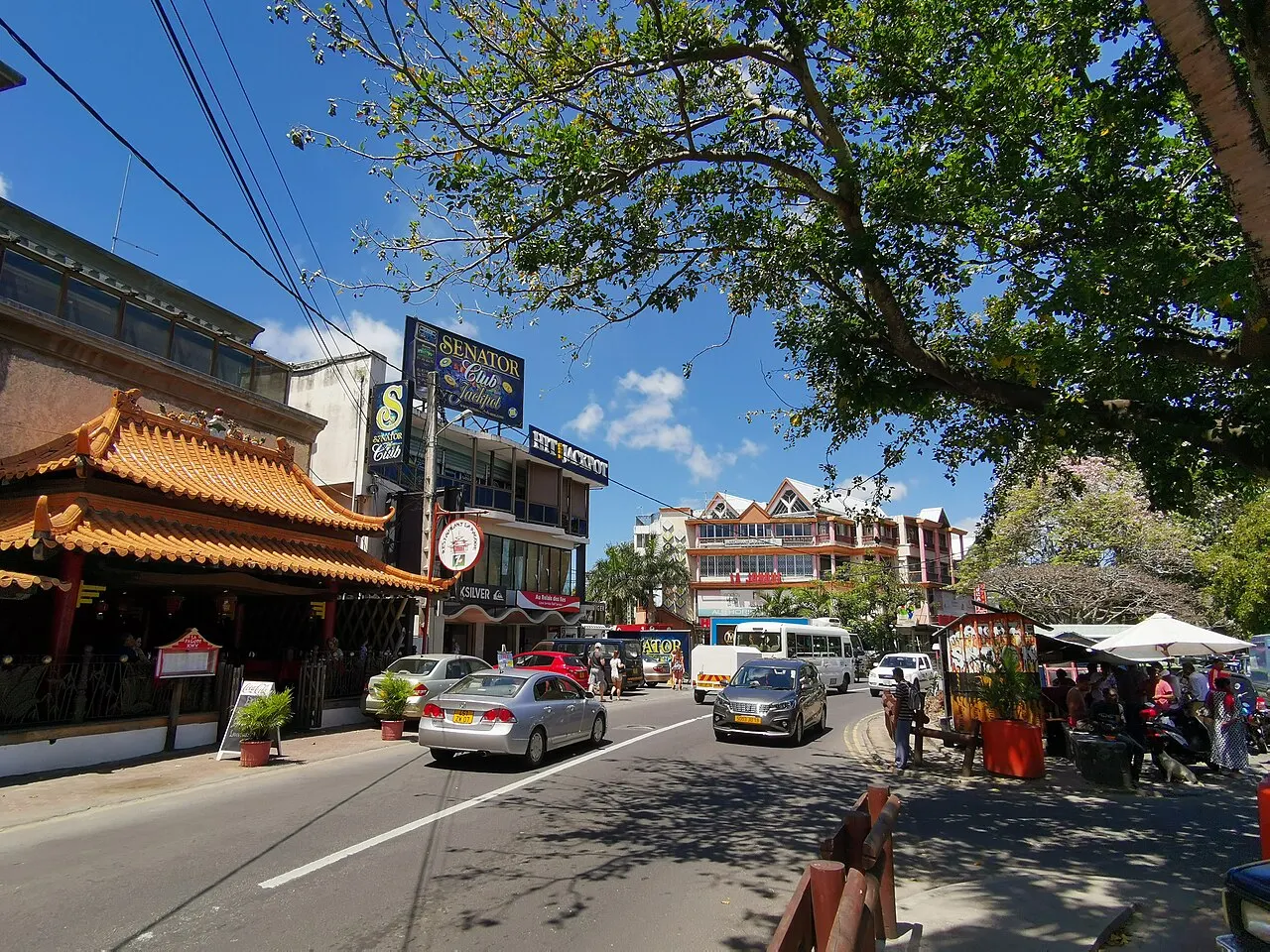 A rua principal de Grand Baie, com restaurantes, uma fachada de telhado chinês, carros e árvores na calçada.