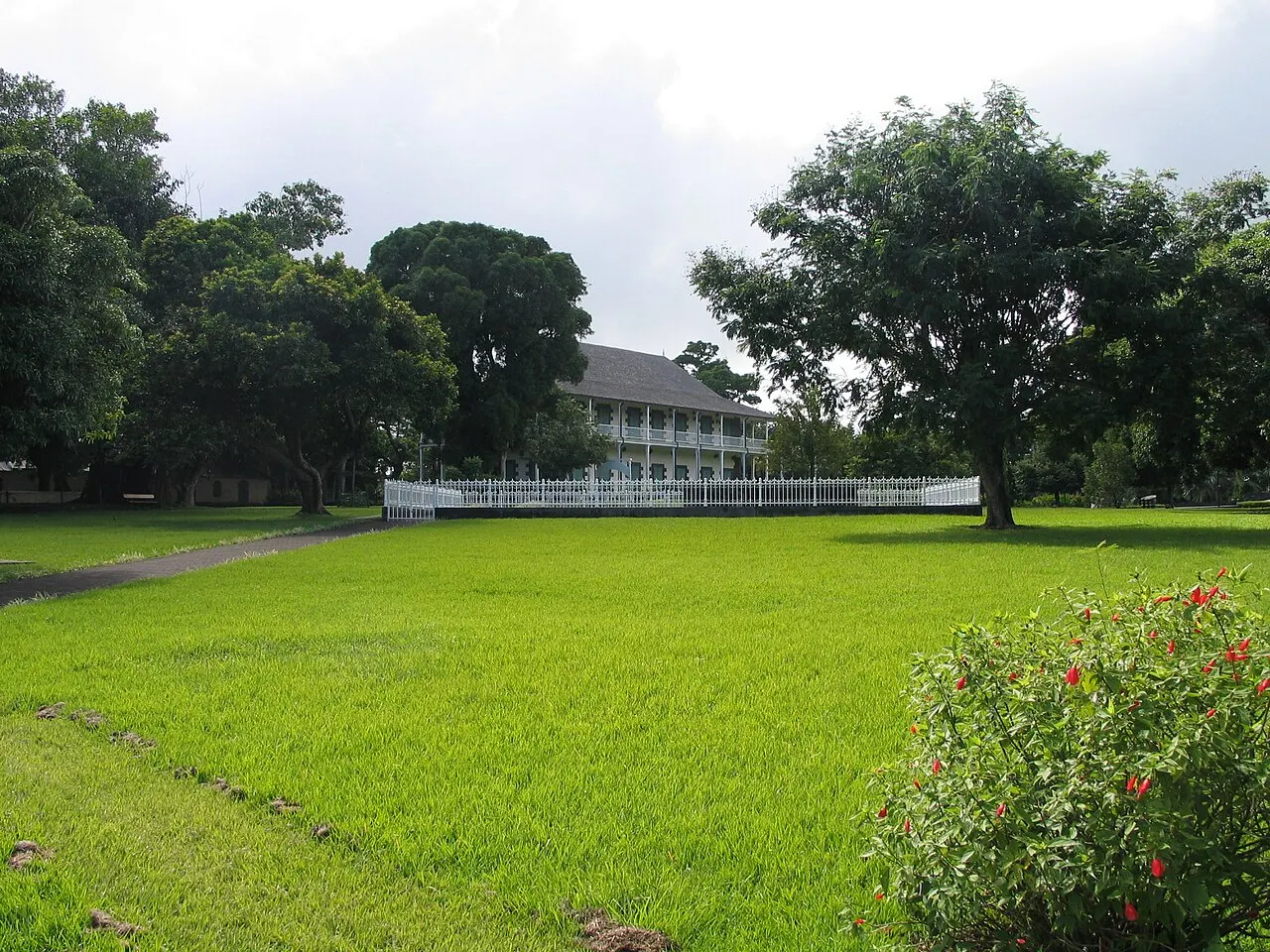 O gramado amplo do jardim botânico de Pamplemousses, com árvores grandes e o casarão branco de Mon Plaisir ao fundo.