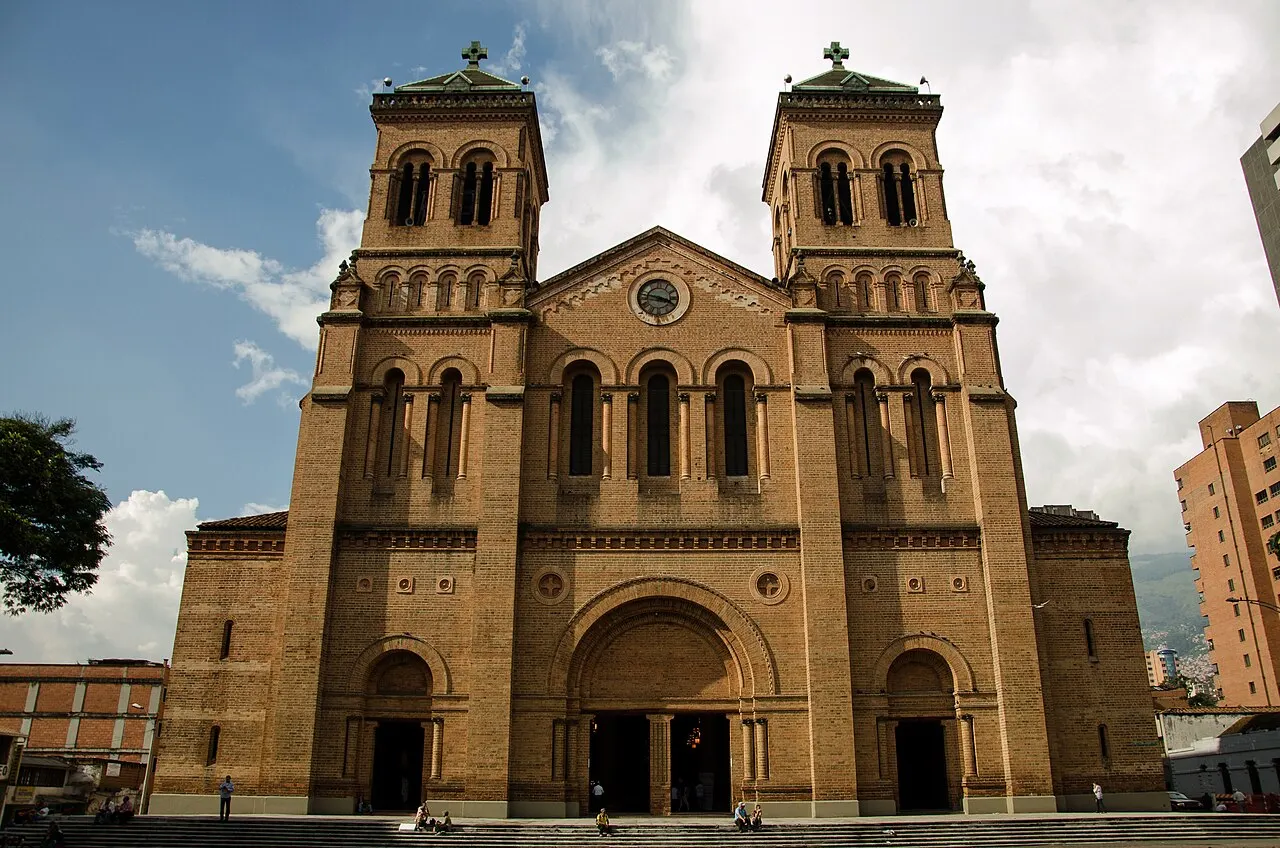Fachada de tijolo aparente da Catedral Metropolitana de Medellín, com as duas torres, sob céu com nuvens.