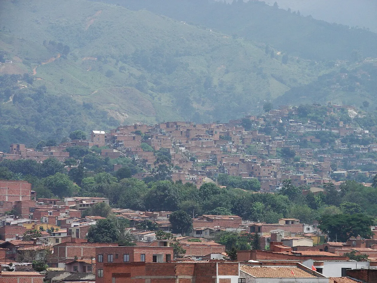 Casas de tijolo aparente e telha vermelha da Comuna 13 cobrindo a encosta de Medellín em camadas, com árvores entre os quarteirões e a serra verde e enevoada fechando o vale ao fundo.