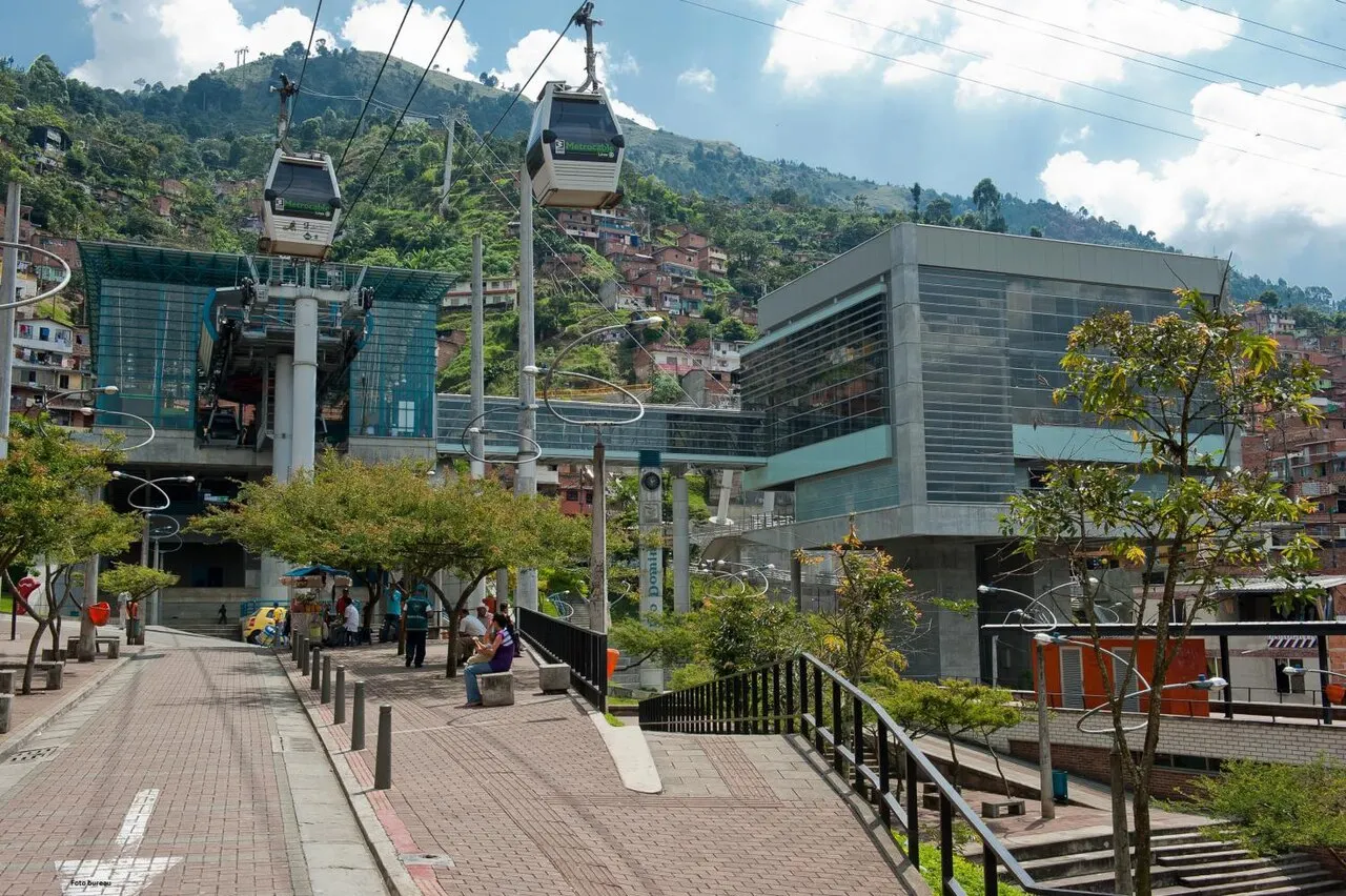 Duas cabines do teleférico Metrocable K suspensas sobre casas na encosta, com a estação de vidro e concreto abaixo, sob céu com nuvens.