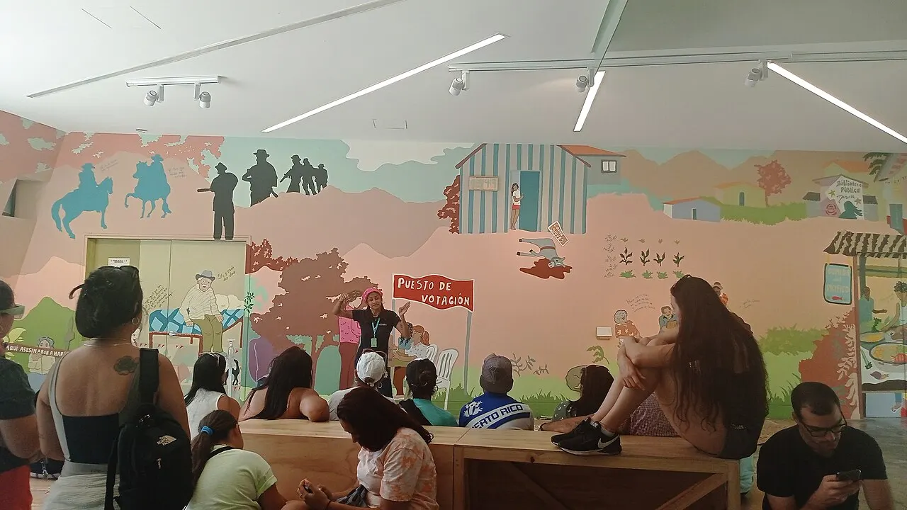 Sala do museu com um grande mural ilustrado cobrindo a parede, mostrando cenas de violência e vida rural, com visitantes sentados em um banco de madeira.