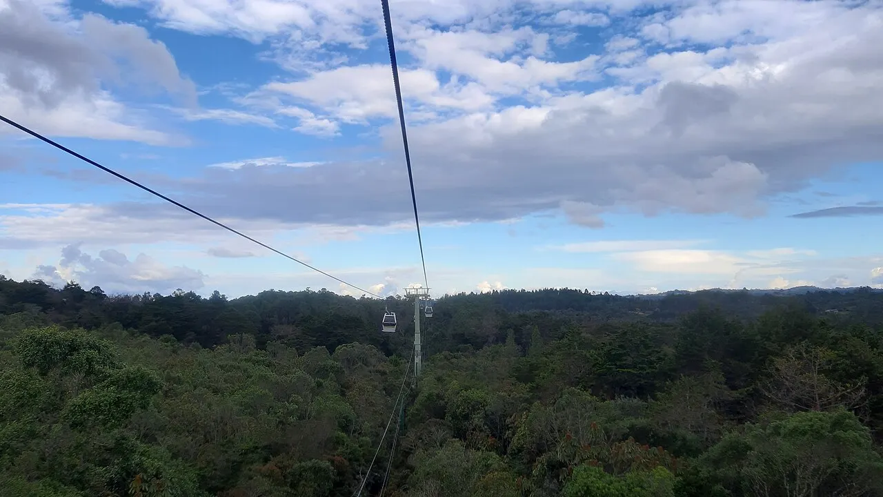 Cabos e cabine do metrocable sobre a mata do Parque Arví, com céu azul e nuvens ao fundo.