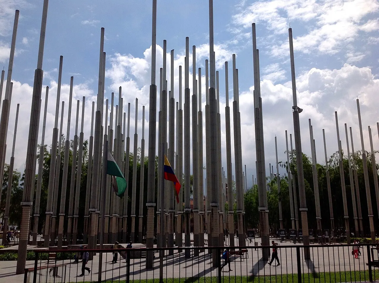 Os postes de luz da Plaza Cisneros, em fileiras densas, sob céu azul com nuvens, no centro de Medellín.