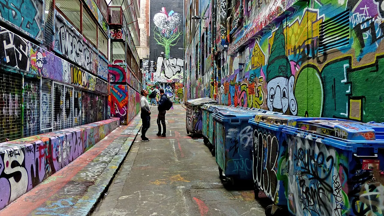 Um beco estreito com as paredes inteiramente cobertas de grafites coloridos, latões de lixo pichados de um lado e duas pessoas conversando no meio do corredor.