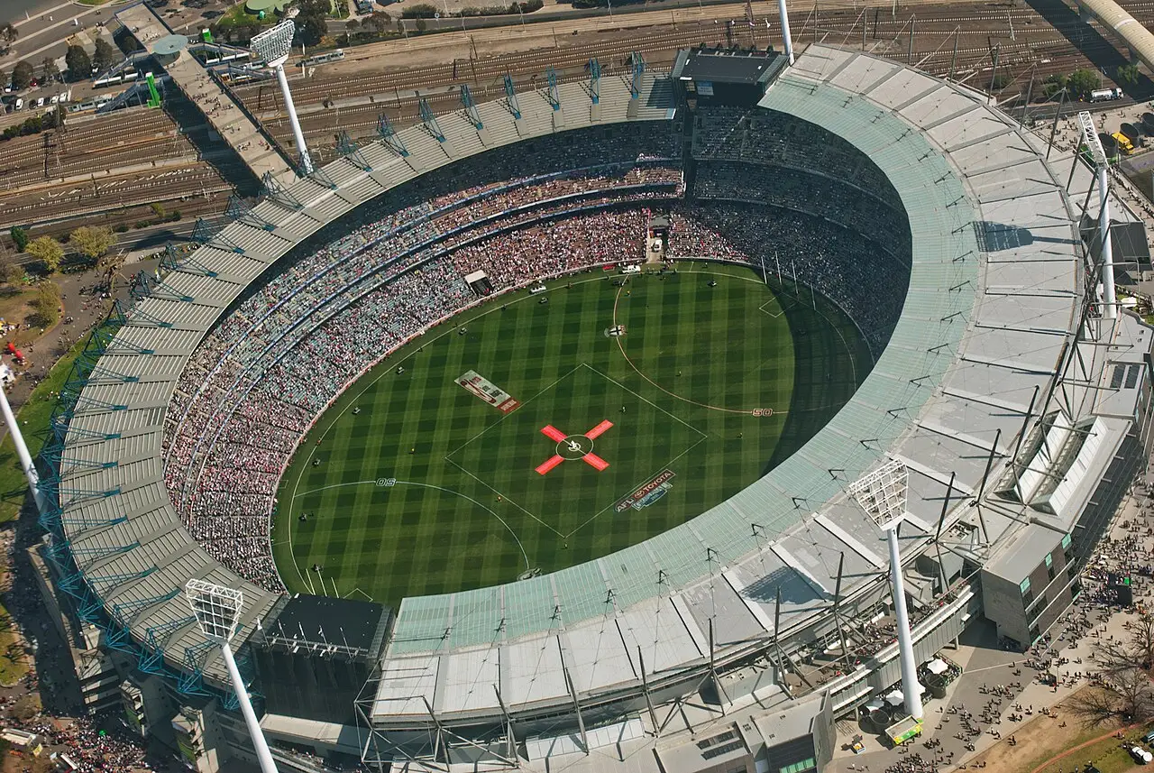 Vista aérea do Melbourne Cricket Ground, com o gramado oval, marcas no centro e as arquibancadas cheias.