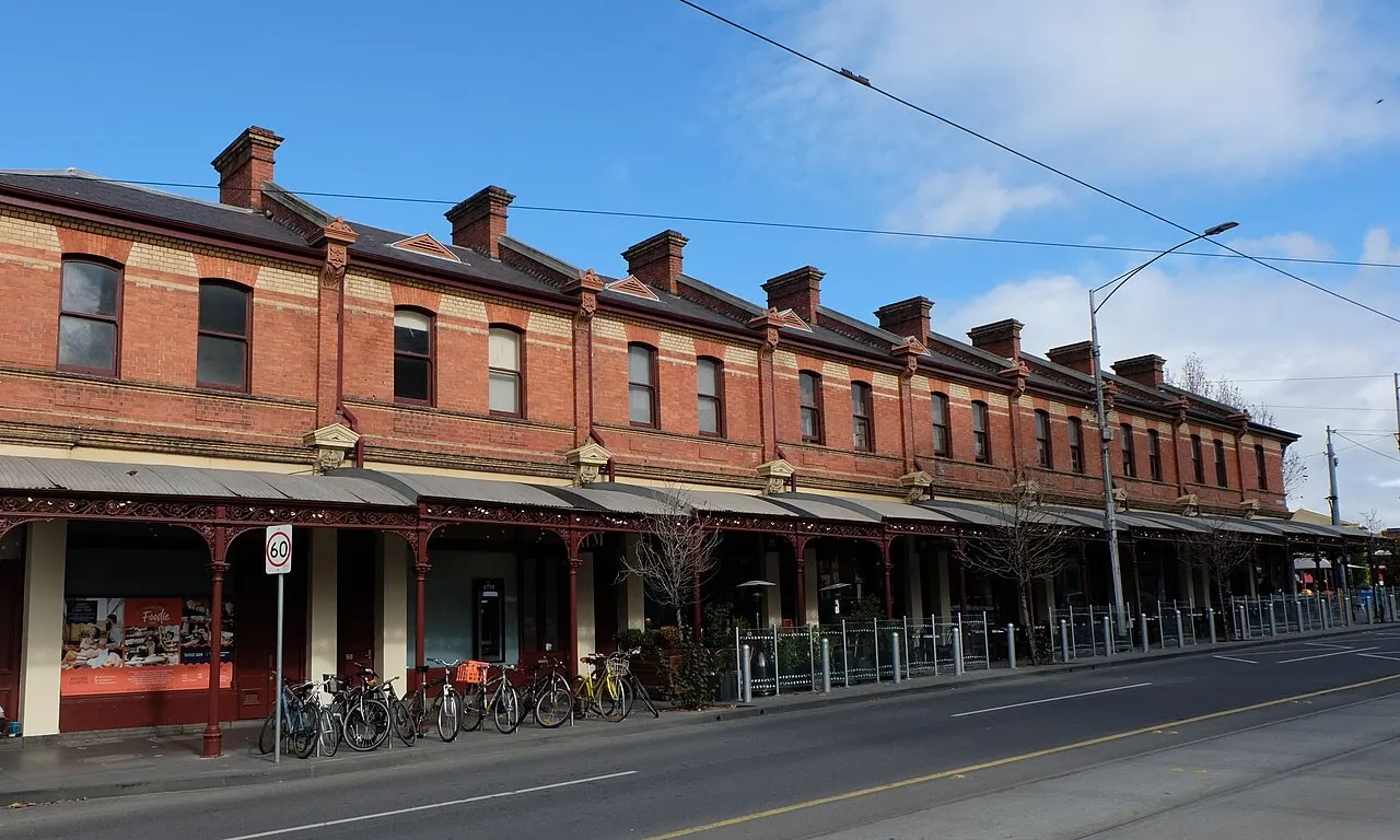 Fileira de lojas em tijolo vitoriano do Queen Victoria Market, com chaminés no telhado, toldos de ferro e bicicletas estacionadas na calçada.