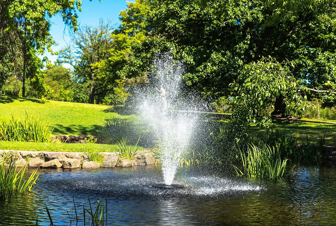 Lago com chafariz nos jardins botânicos de Melbourne, cercado por gramado e árvores em um dia de sol.