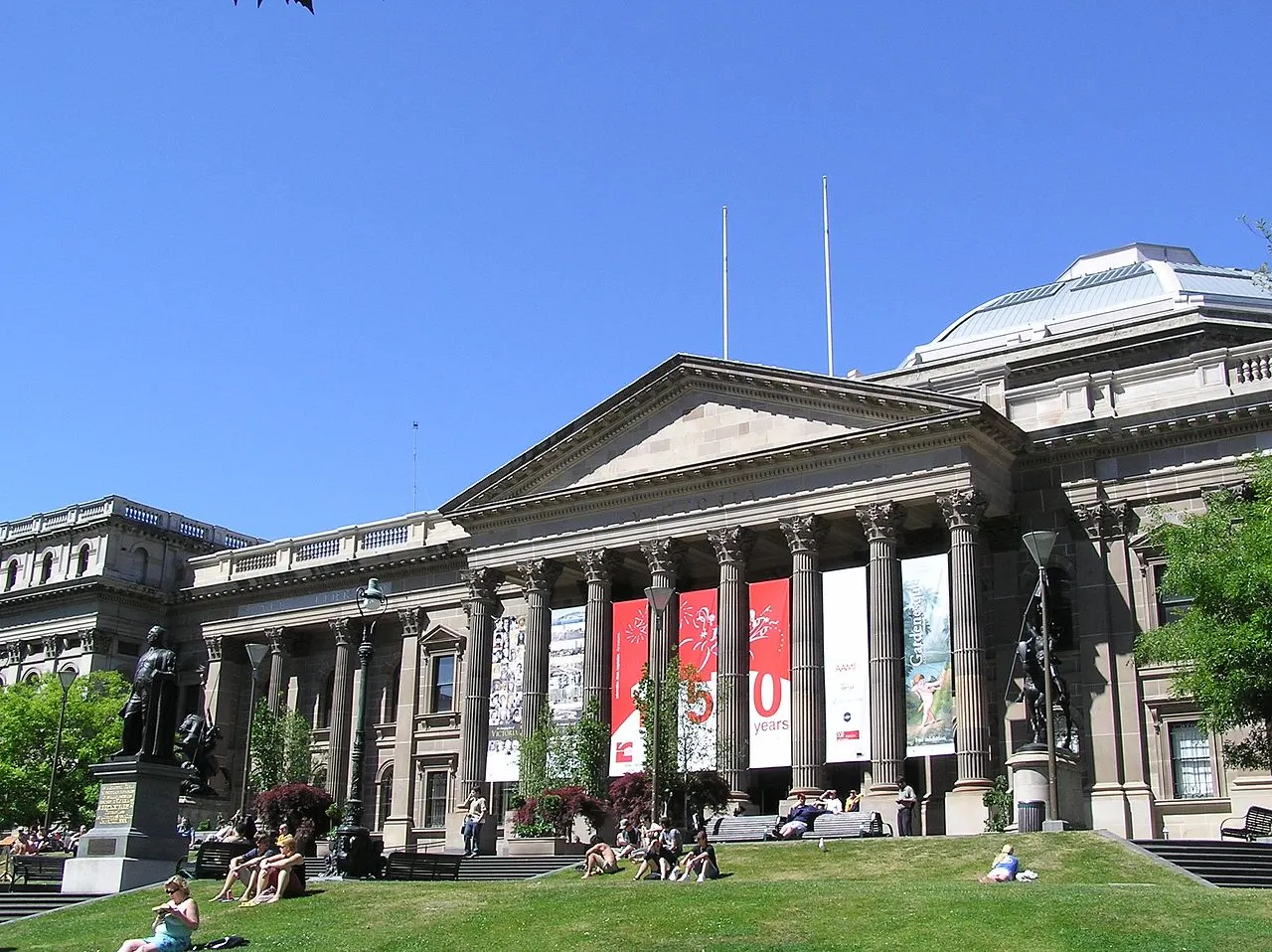 Fachada neoclássica da State Library Victoria, com pórtico de colunas coríntias, frontão triangular e pessoas sentadas no gramado em frente, sob céu azul.
