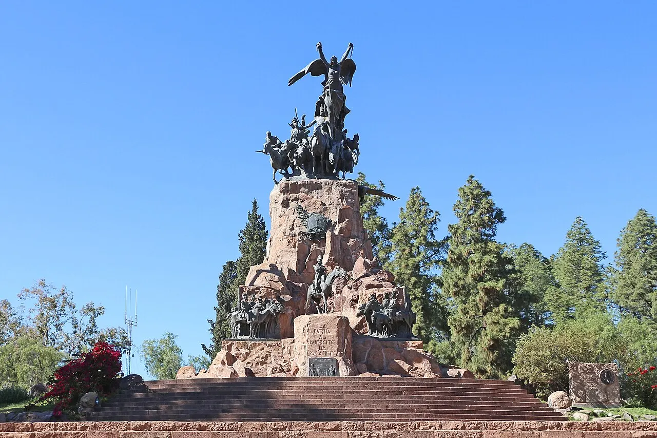 O Monumento ao Exército dos Andes, no Cerro de la Gloria, com grupos de figuras de bronze sobre uma base de pedra avermelhada, entre pinheiros, sob céu azul.