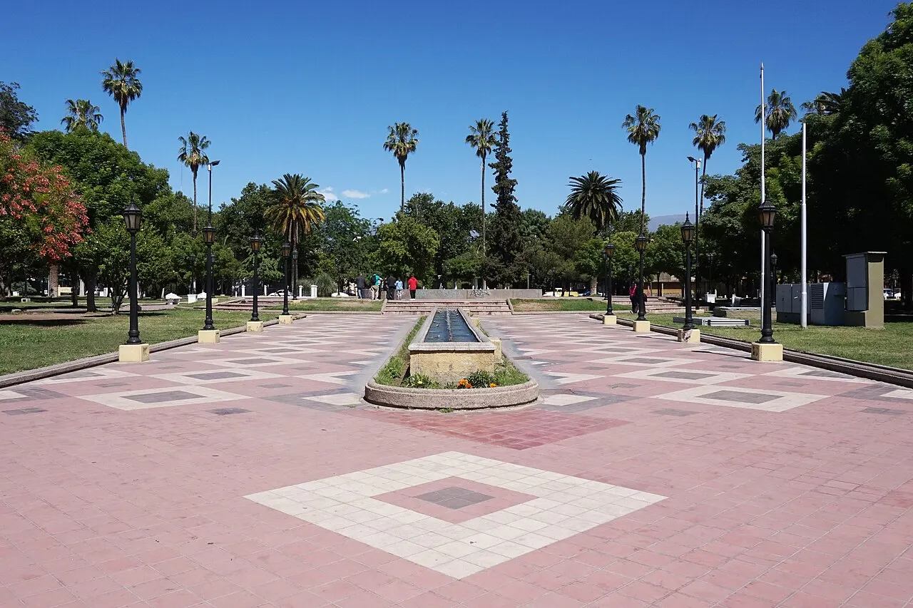 A Plaza Pedro del Castillo, na Área Fundacional de Mendoza, com piso rosado de losangos, um espelho d'água baixo no centro e palmeiras ao redor, sob céu azul.
