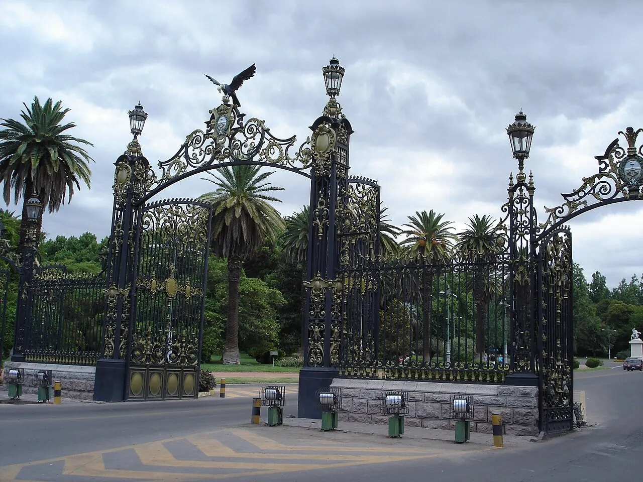 Os portões de ferro ornamentado do Parque General San Martín, em Mendoza, com um condor de metal no topo, postes de luz antigos e palmeiras atrás.