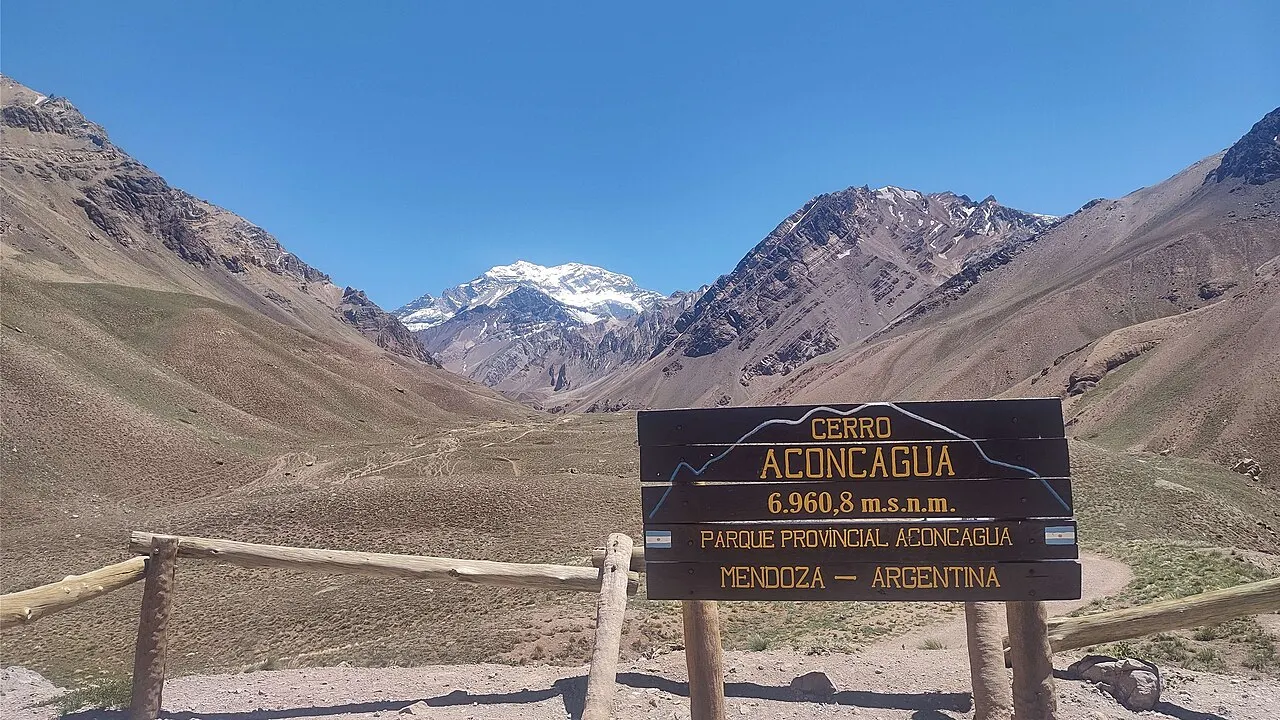 Uma placa de madeira com os dizeres «Cerro Aconcagua 6.960,8 m s.n.m.» diante de um vale seco, com o pico nevado do Aconcágua ao fundo, entre montanhas marrons.