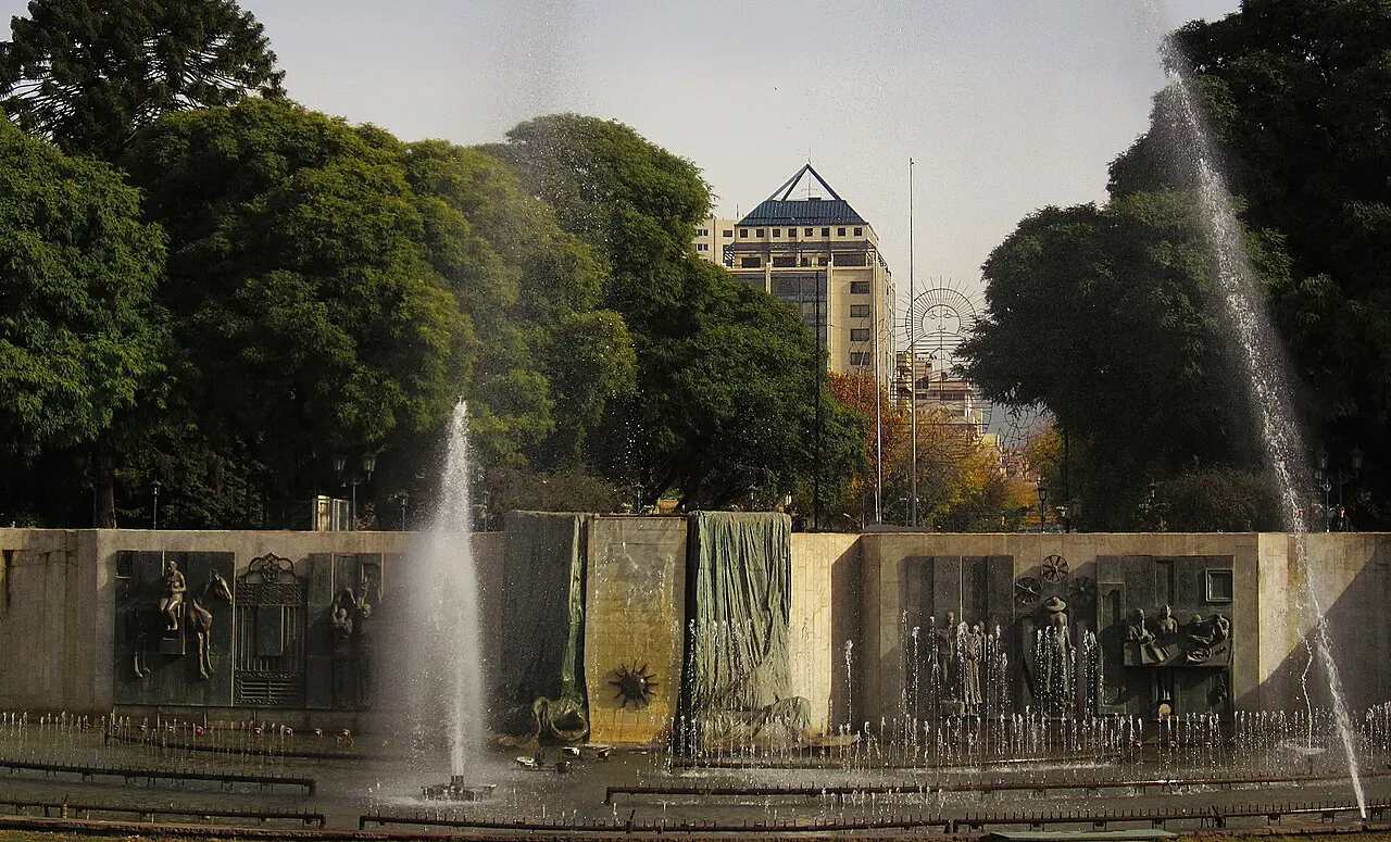 A fonte da Plaza Independencia, em Mendoza, com jatos d'água altos diante de um muro de painéis em relevo, árvores ao redor e um prédio alto ao fundo.