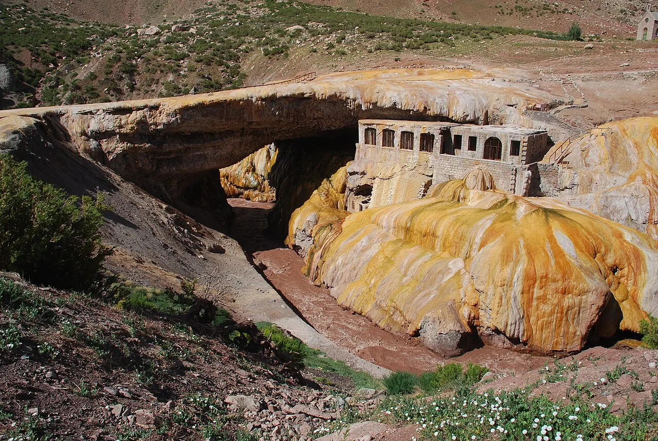 O Puente del Inca, um arco natural de pedra sobre o rio, com depósitos minerais alaranjados e amarelos e as ruínas do antigo balneário encostadas na rocha.