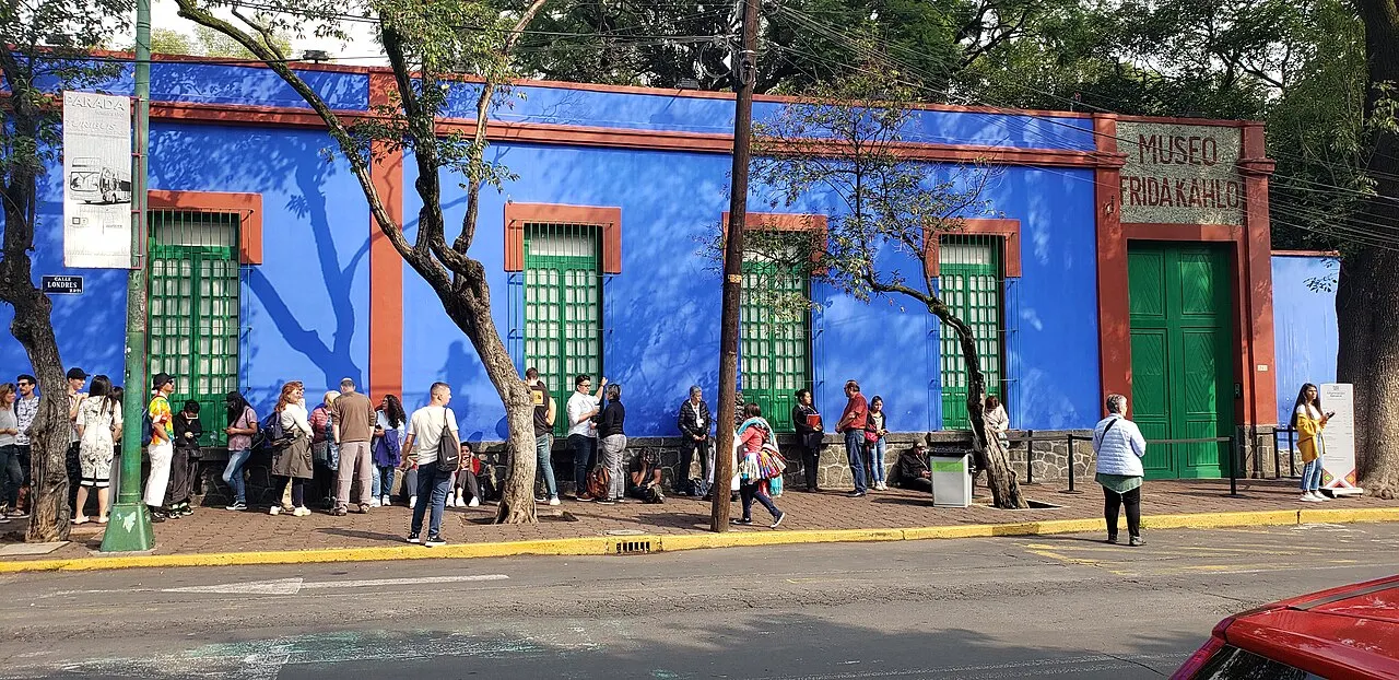 Muro azul-cobalto do Museu Frida Kahlo com portas e janelas verdes e faixa vermelha no alto; na calçada em frente, uma fila de visitantes à sombra das árvores.