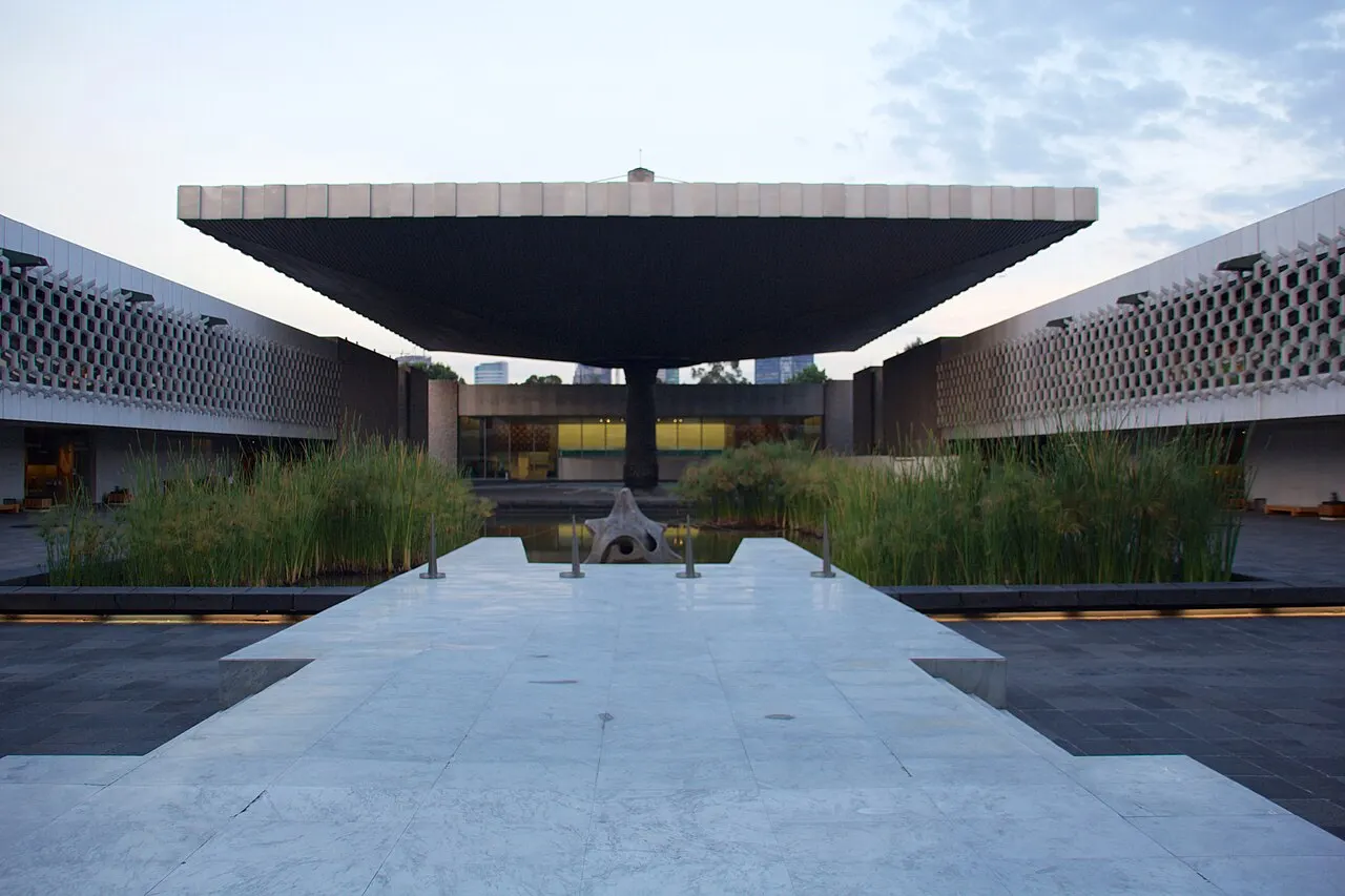 Grande cobertura retangular suspensa por uma única coluna central forma o teto do pátio do museu, sobre um espelho d'água com vegetação e prédios ao fundo.