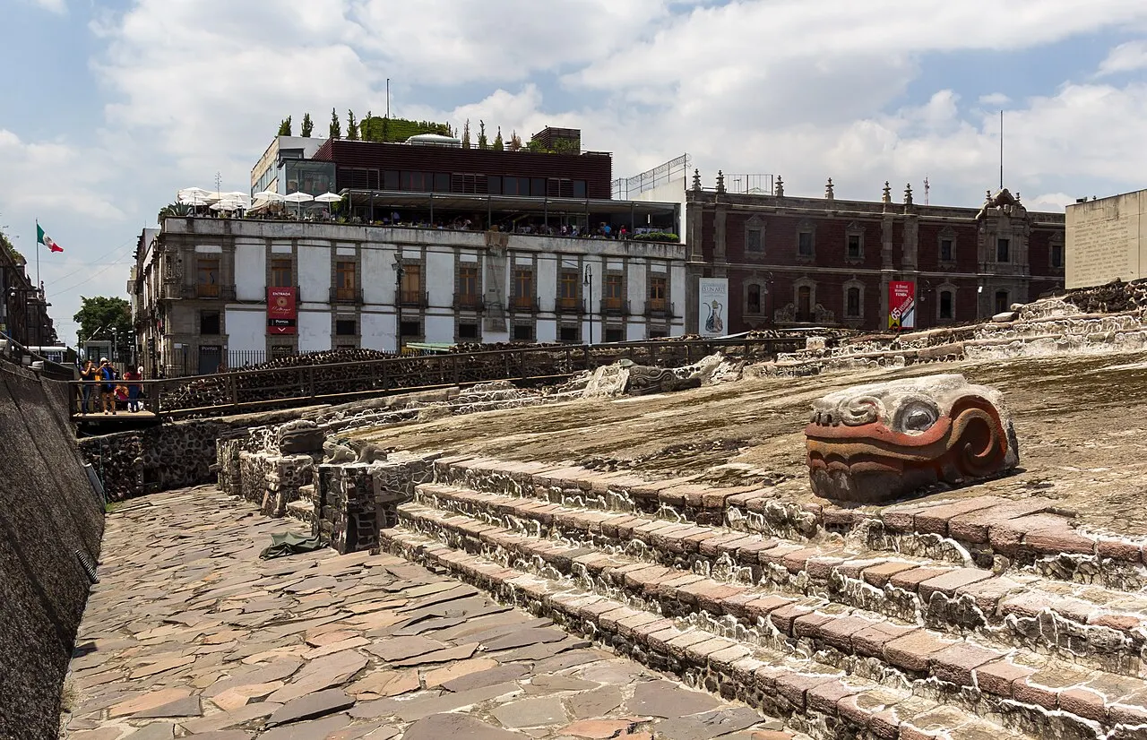 Ruínas de pedra em degraus do Templo Mayor, com uma cabeça de serpente esculpida em primeiro plano e prédios coloniais ao fundo, sob céu com nuvens.