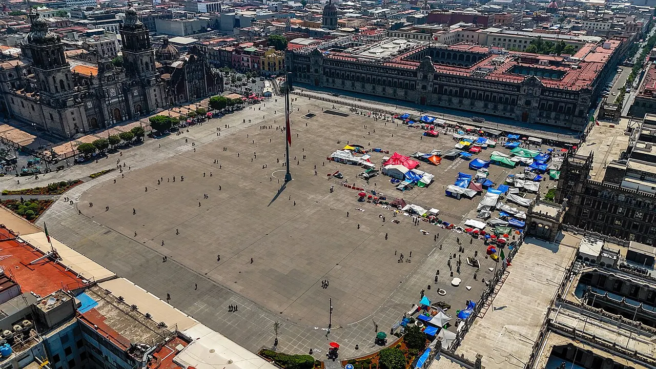 Vista aérea do Zócalo da Cidade do México, com a praça ampla, barracas coloridas e a catedral à esquerda.