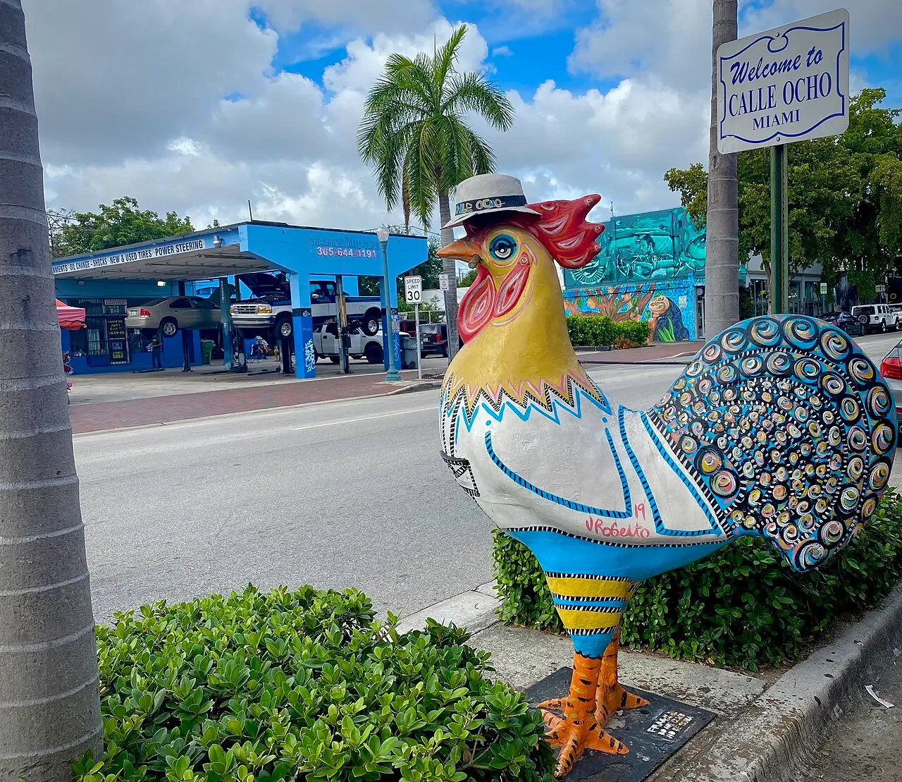 Estatua colorida de un gallo con sombrero, pintada con patrones geométricos, junto a un cartel de bienvenida a la Calle Ocho, con una palmera y un taller al fondo.