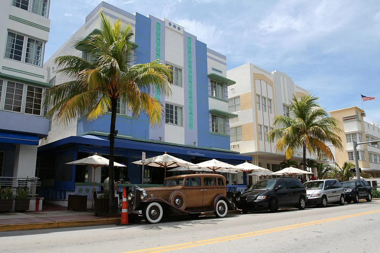 Prédios Art Déco pintados de azul e branco na Ocean Drive, com palmeiras, um carro antigo estacionado e mesas com guarda-sóis na calçada, sob céu azul.