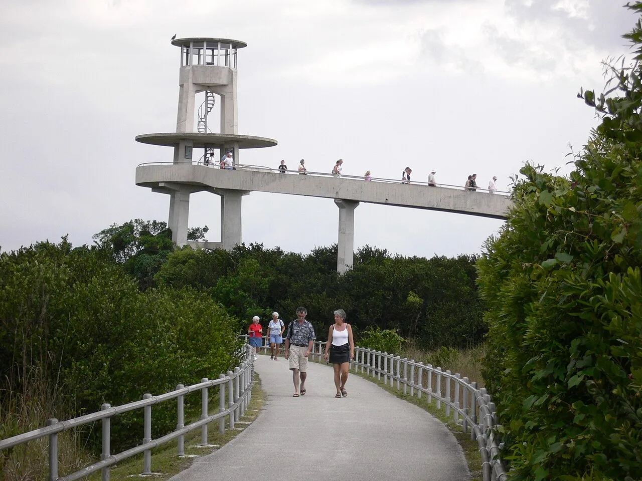 Torre de concreto com rampa elevada em curva, cercada de mata baixa, e visitantes caminhando pela passarela que leva até ela.