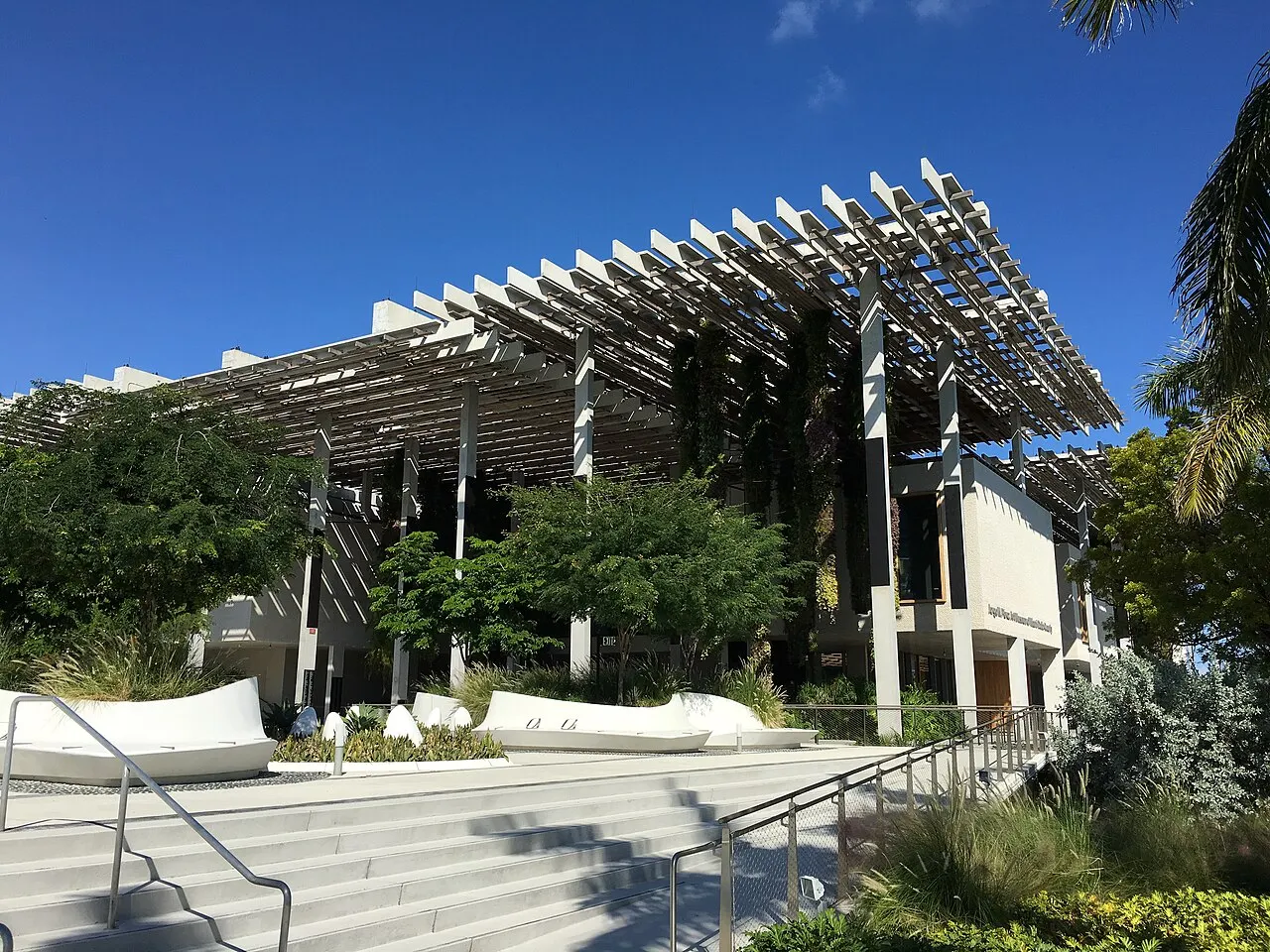 Fachada del Pérez Art Museum Miami, con una cubierta calada de listones suspendida sobre columnas altas, plantas colgantes y bancos blancos curvos en el jardín.