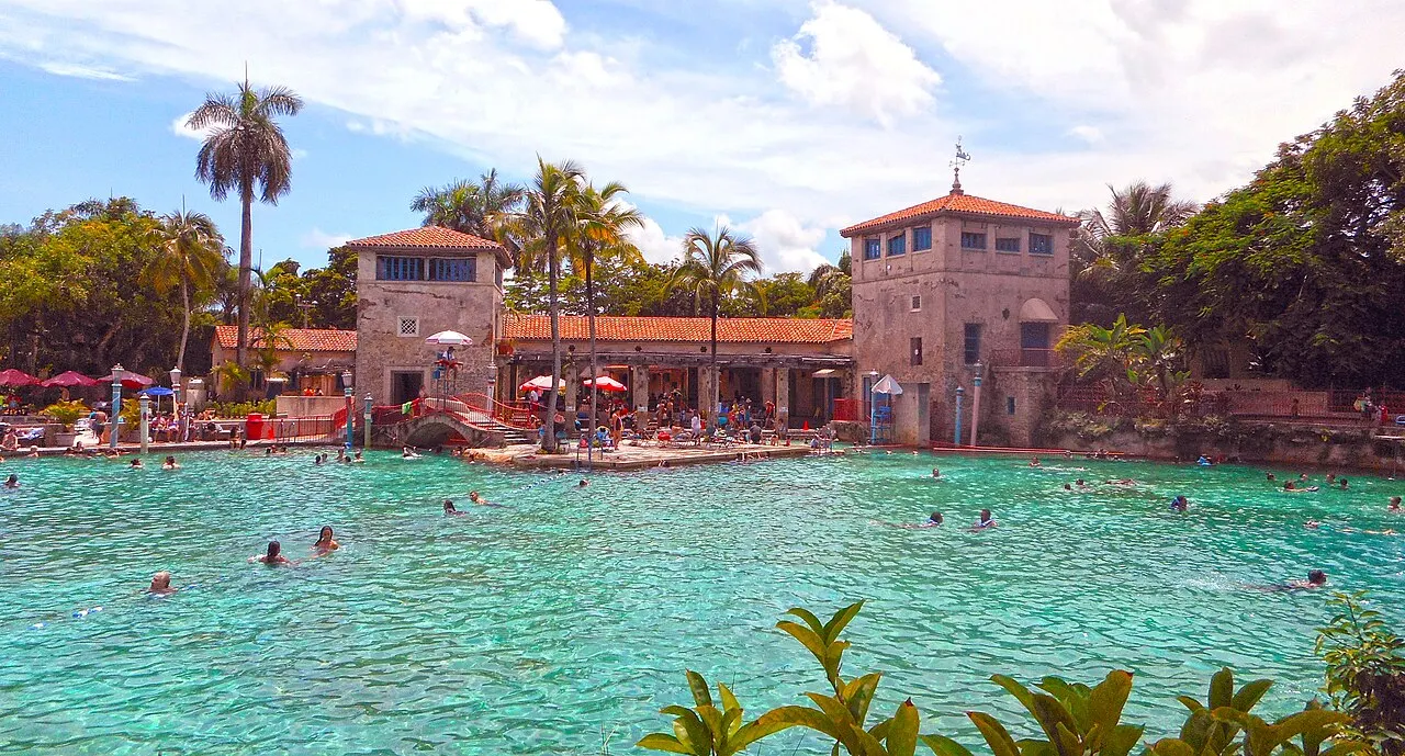 A Venetian Pool, com água turquesa, banhistas e torres de estilo mediterrâneo cercadas por palmeiras.