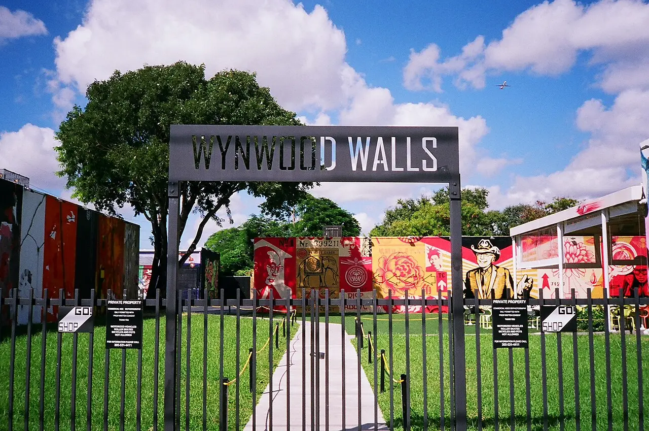 Entrada do Wynwood Walls, com o nome no pórtico preto, o gramado e murais coloridos atrás da grade.