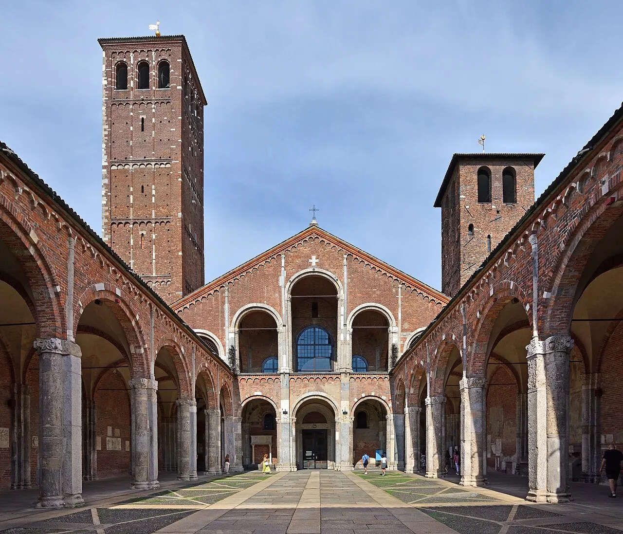 Átrio de arcos de tijolo da Basilica di Sant’Ambrogio, com a fachada e as duas torres ao fundo.