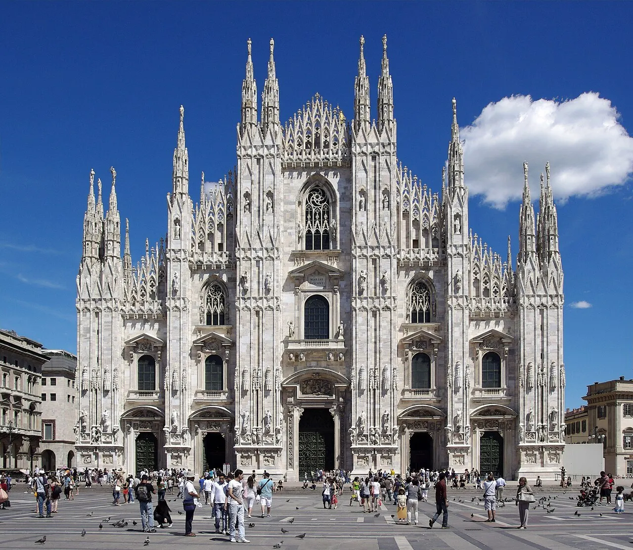 Fachada de mármore branco do Duomo de Milão, com pináculos e agulhas, sob céu azul, com a praça cheia embaixo.