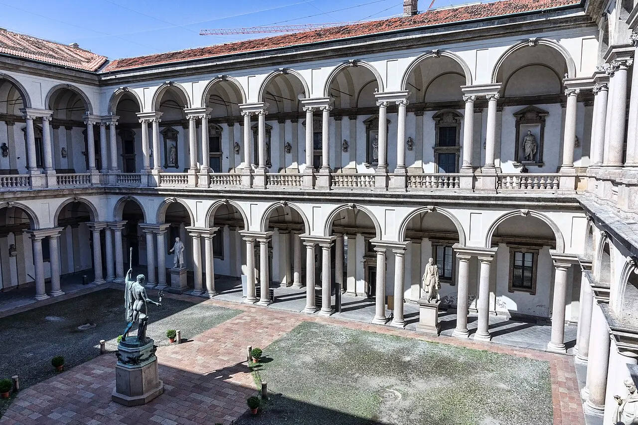 Pátio interno da Pinacoteca di Brera, com duas galerias de arcos e colunas sobrepostas e uma estátua de bronze ao centro, sob céu azul.
