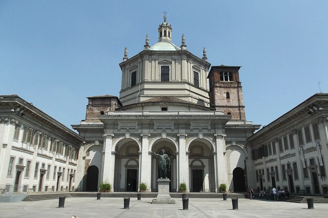 Fachada da Basilica di San Lorenzo Maggiore, com o pórtico de colunas e a cúpula ao fundo, sob céu azul.