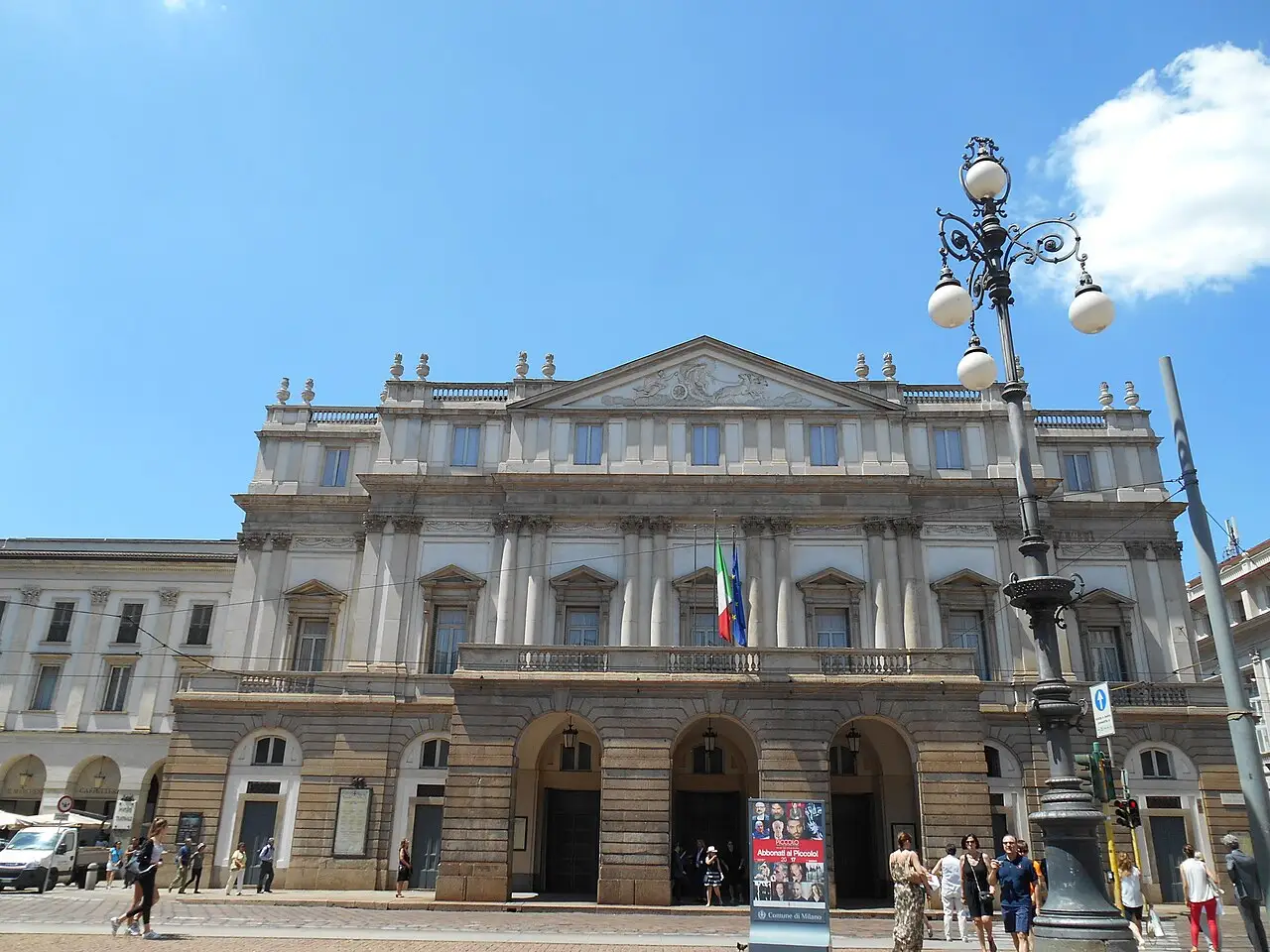 Fachada do Teatro alla Scala, com frontão, arcos no térreo, a bandeira italiana na sacada e um poste na calçada.