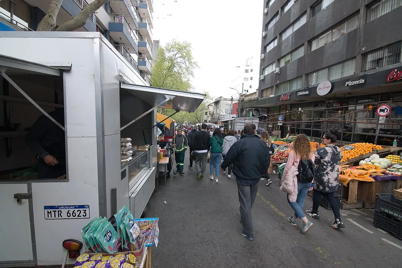 Rua Tristán Narvaja tomada pela feira: um trailer de venda aberto à esquerda, bancas de laranja, abóbora e verdura à direita, caixas empilhadas na calçada e gente caminhando pelo meio da rua entre prédios altos.