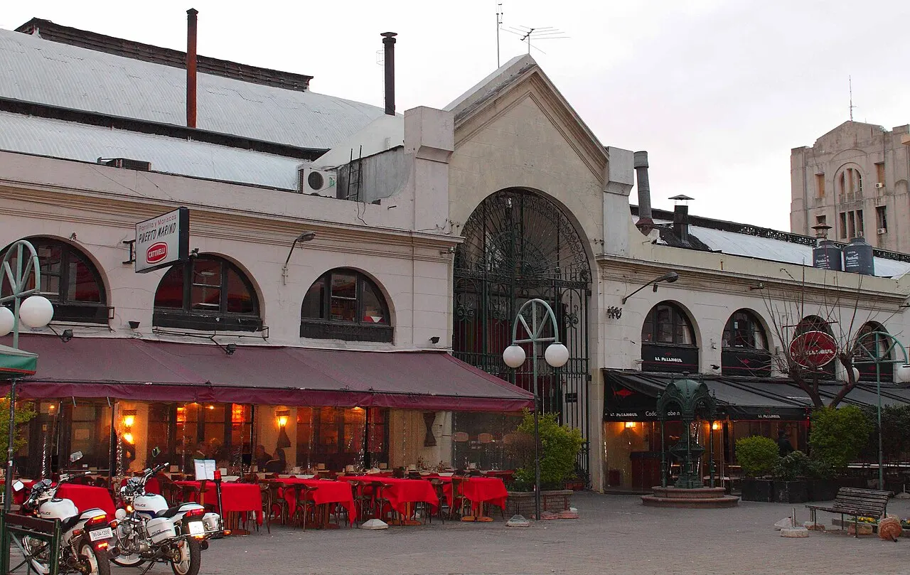 Fachada do Mercado del Puerto ao entardecer, com portão de ferro em arco, mesas de restaurante com toalhas vermelhas na calçada e motos estacionadas em frente.