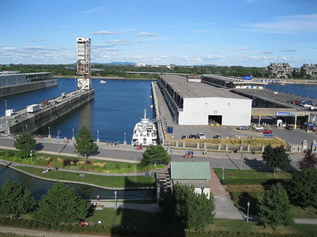 Vista elevada do Vieux-Port de Montreal, com uma torre de ponte levadiça, galpões junto ao cais, um barco atracado e prédios ao fundo sob céu com nuvens.