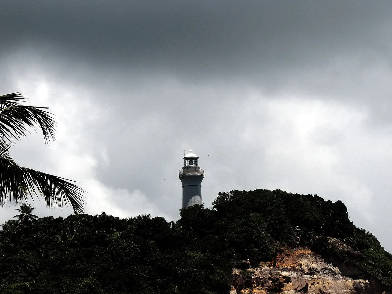 O farol branco no alto do morro, recortado contra um céu de nuvens carregadas, com coqueiros na frente.