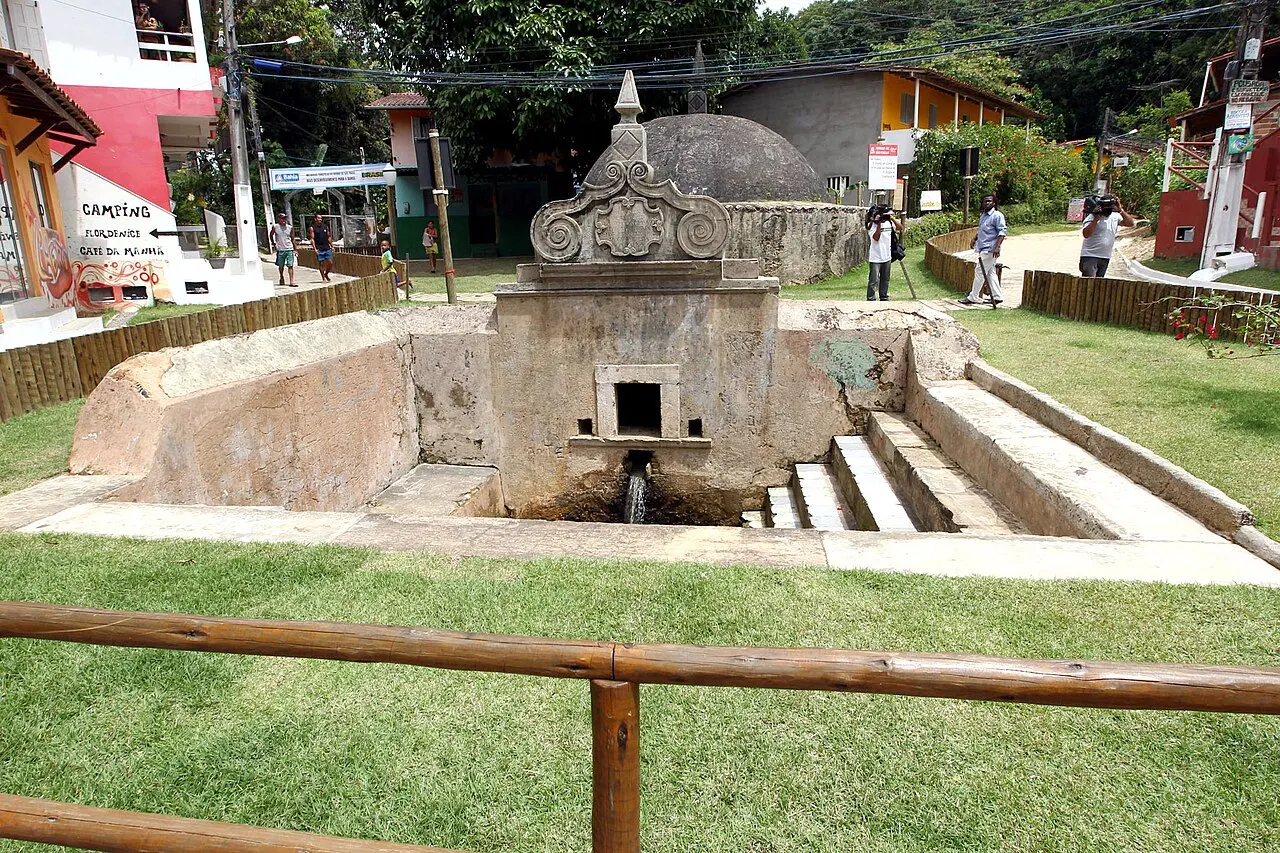 A Fonte Grande, chafariz colonial de pedra com escadas que descem até a bica, no gramado da praça da vila.