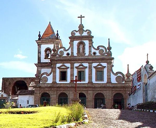 A fachada branca e ornamentada da igreja do Convento de Santo Antônio, em Cairu, com detalhes vermelhos e a cruz no alto.