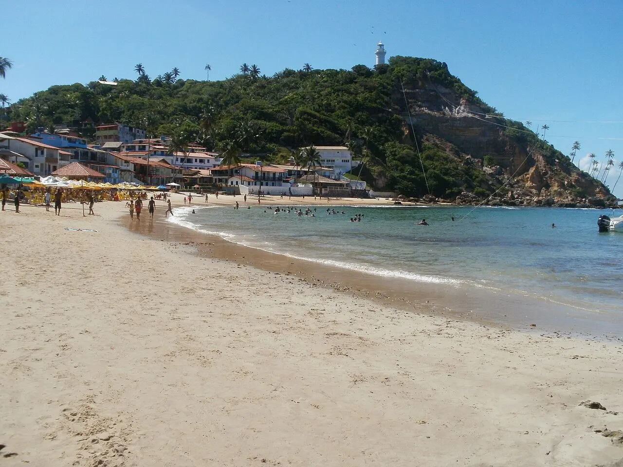 A Primeira Praia, de areia clara, com o morro coberto de mata e as casas da vila no alto, ao fundo.