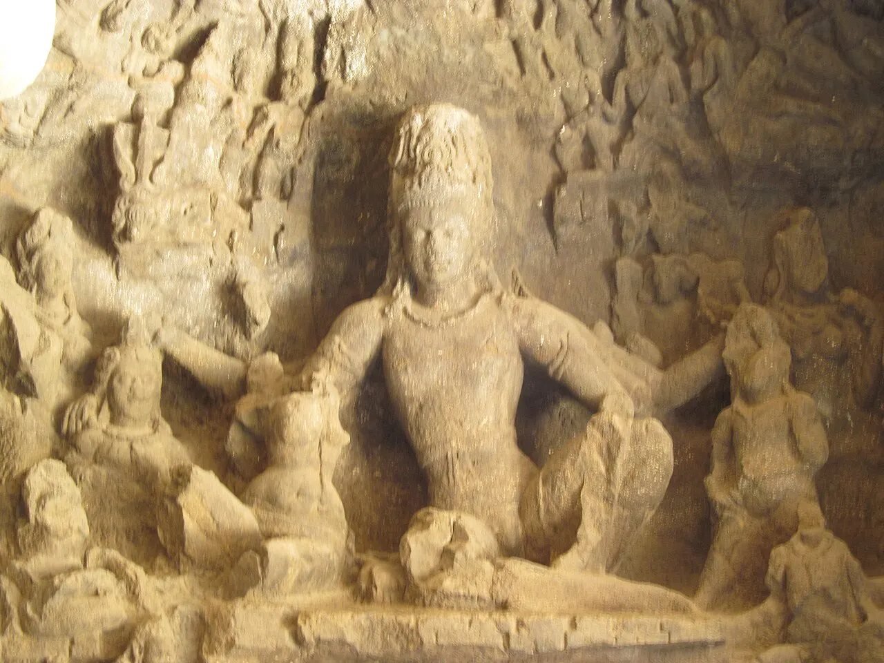 Figura sentada esculpida em pedra clara, muito desgastada pelo tempo, contra fundo escuro da caverna.