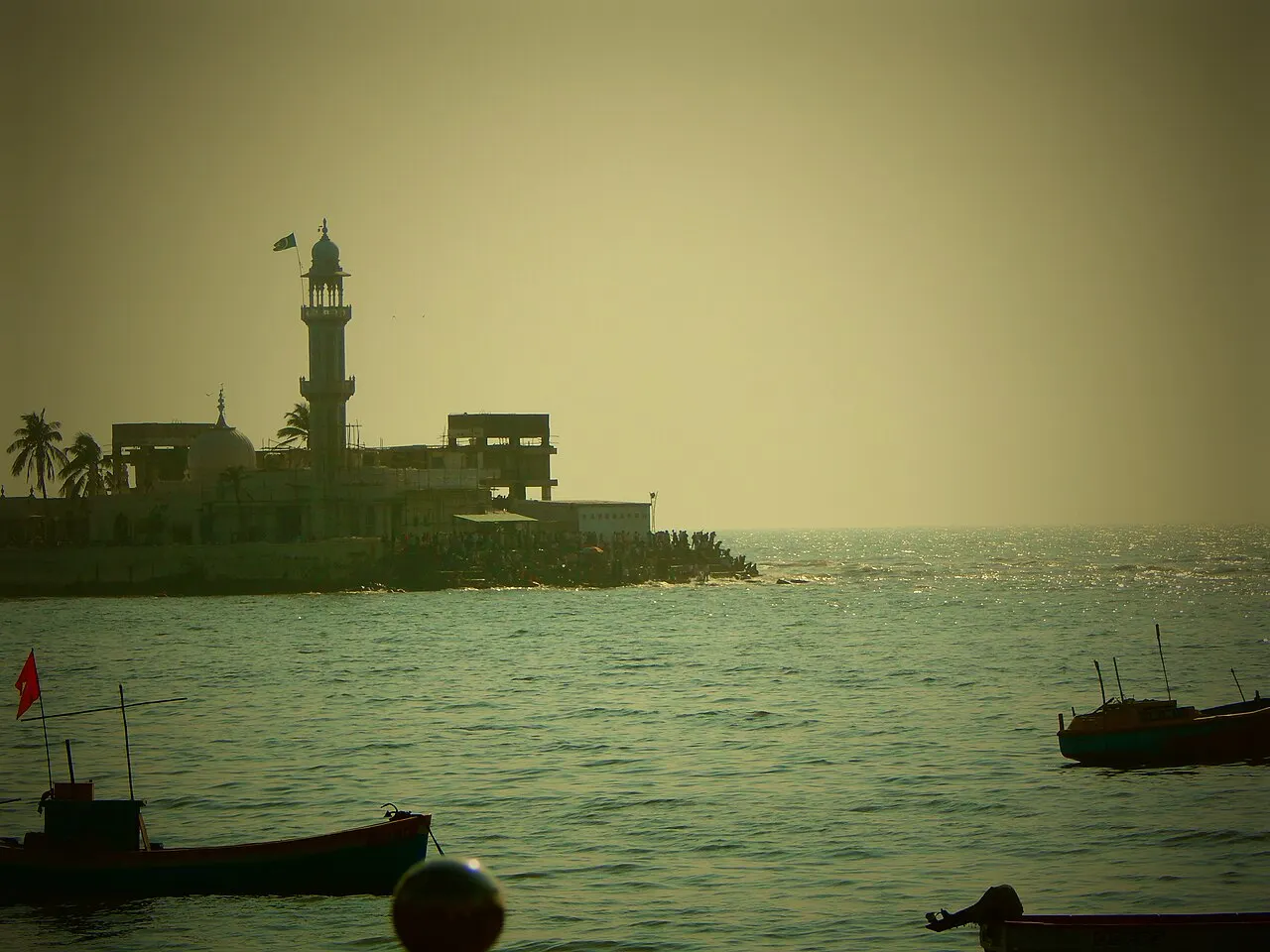 O santuário de Haji Ali recortado em contraluz no fim do quebra-mar que entra no mar da Arábia, com fiéis na passagem estreita e barcos de pesca em primeiro plano.