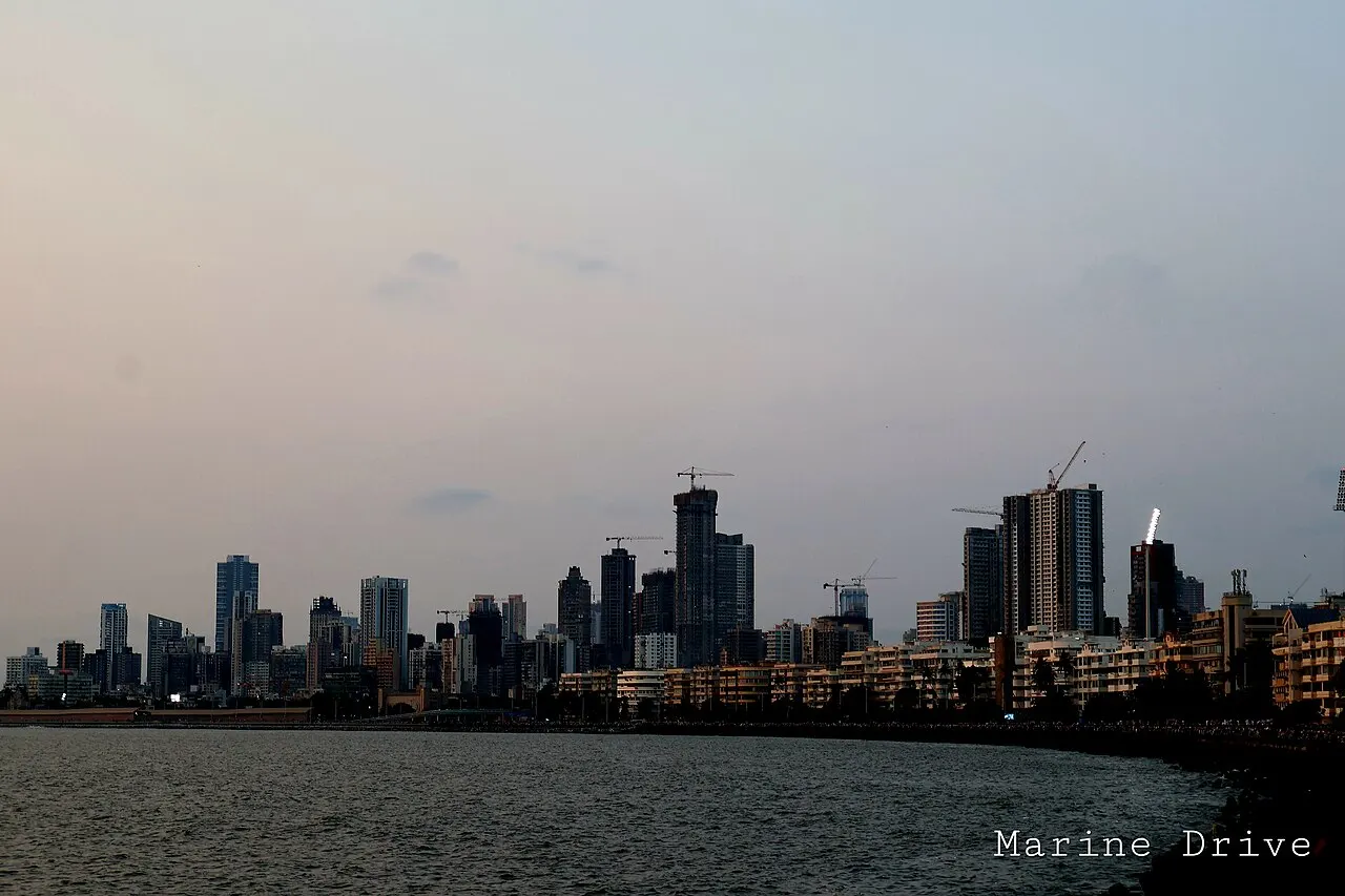 Horizonte de arranha-céus e prédios ao entardecer, visto do outro lado da baía, com guindastes de construção entre as torres e a orla iluminada na base.