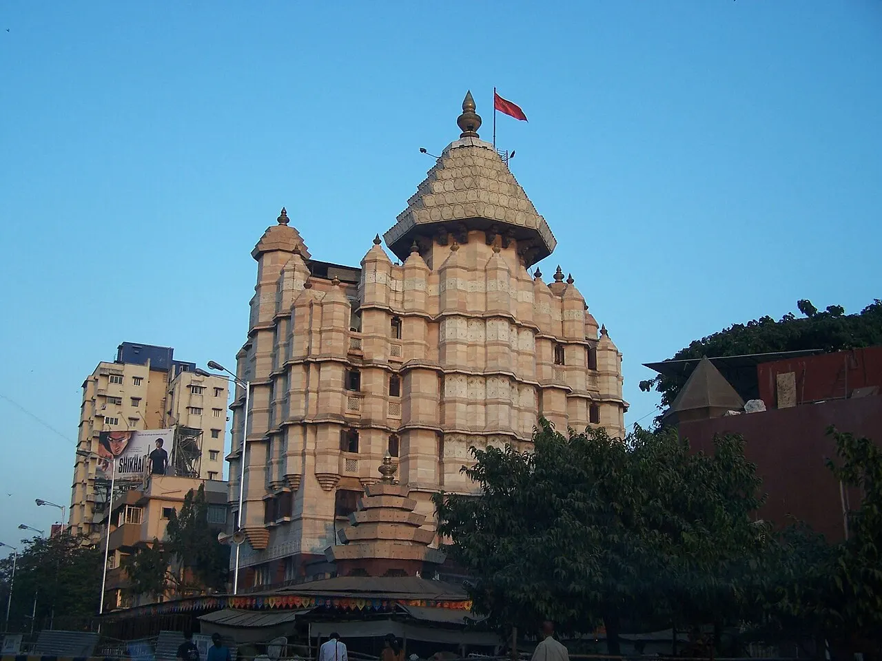 Torre de templo hindu com cúpula dourada e relevos esculpidos, entre prédios residenciais.