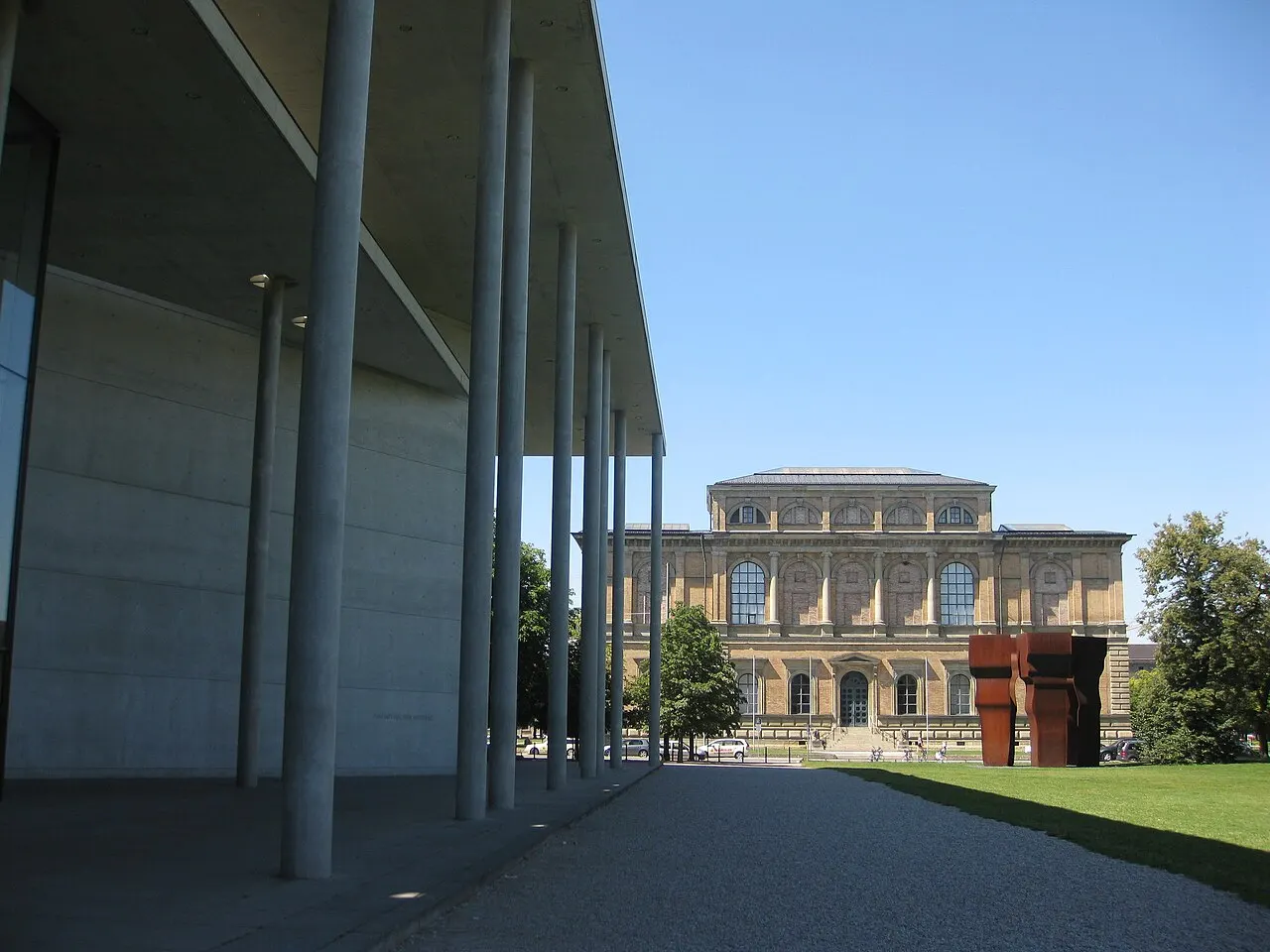 Fachada da Alte Pinakothek, em Munique, com janelas em arco, uma árvore e uma escultura de aço à frente.